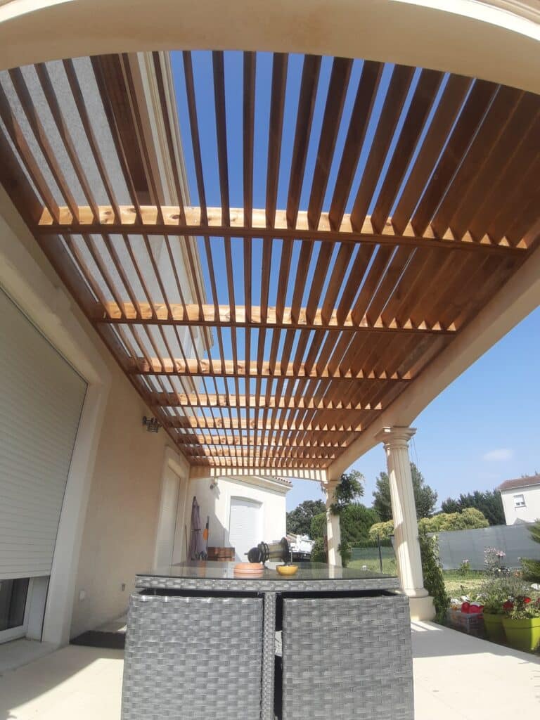 Pergola en bois sur une terrasse avec poteaux à colonnes, vue vers le ciel bleu, au-dessus d'un salon de jardin en résine tressée grise.