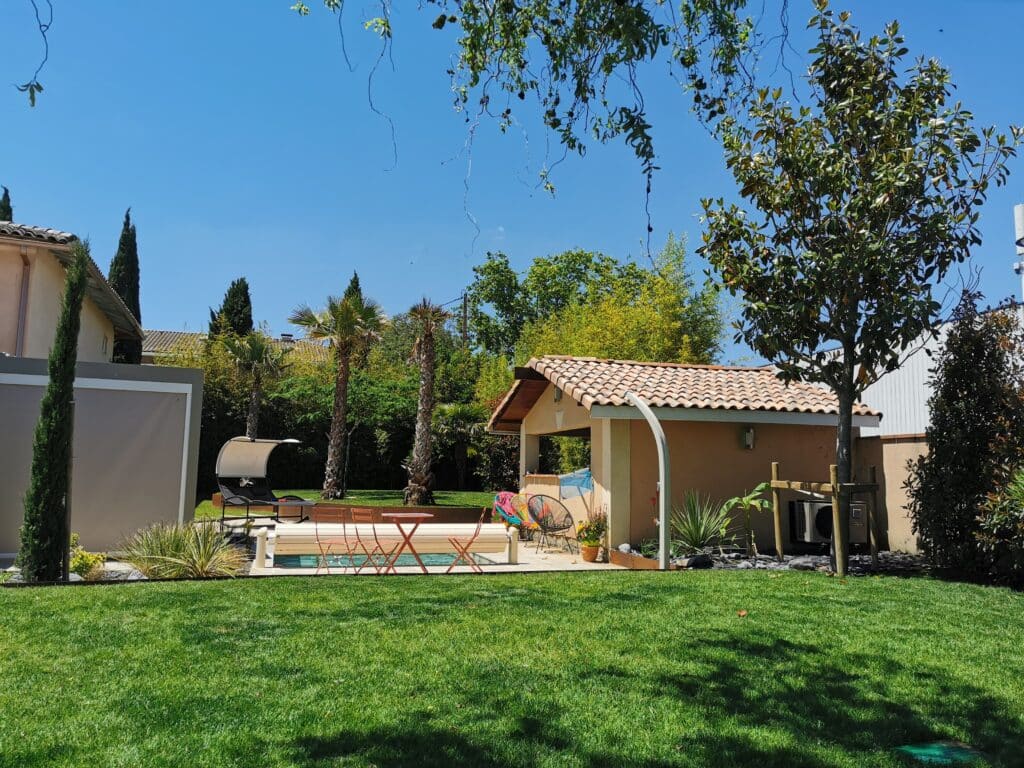 Jardin ensoleillé avec pelouse verte, piscine couverte, pool house à tuiles romaines, palmiers et mobilier de jardin sous un ciel bleu clair.