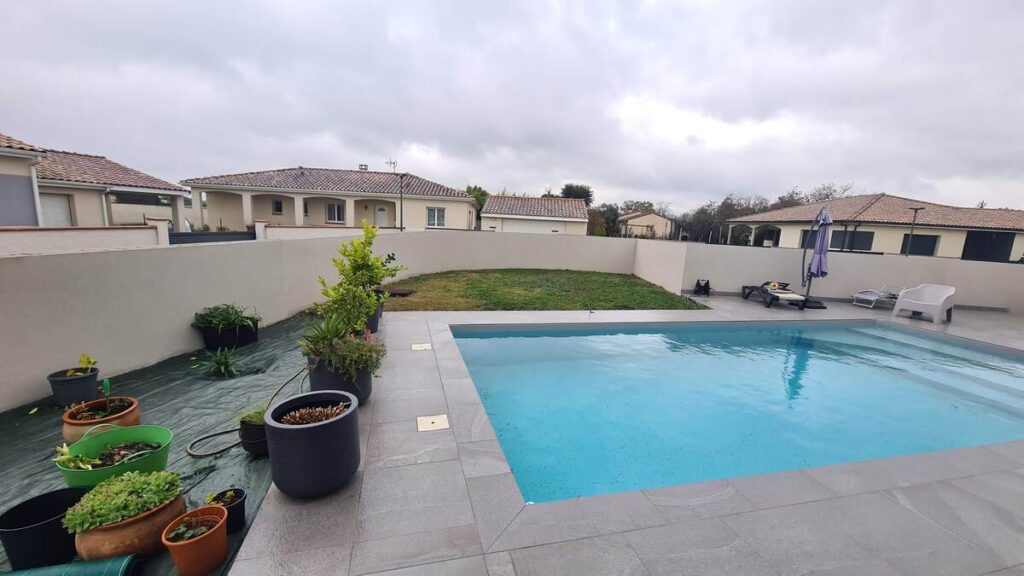 Piscine moderne carrelée avec terrasse et pelouse, entourée d'un mur d'enceinte clair et de plantes en pots.