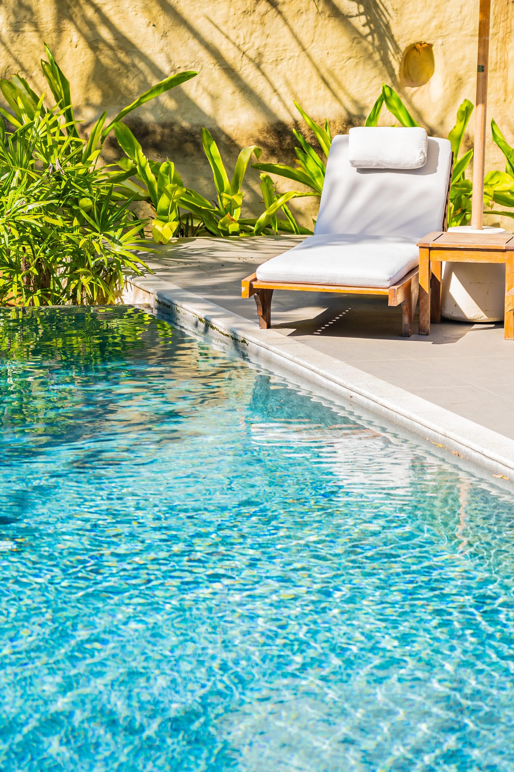 Chaise longue blanche au bord d'une piscine aux eaux turquoise, entourée de verdure luxuriante et d'un mur de couleur sable.