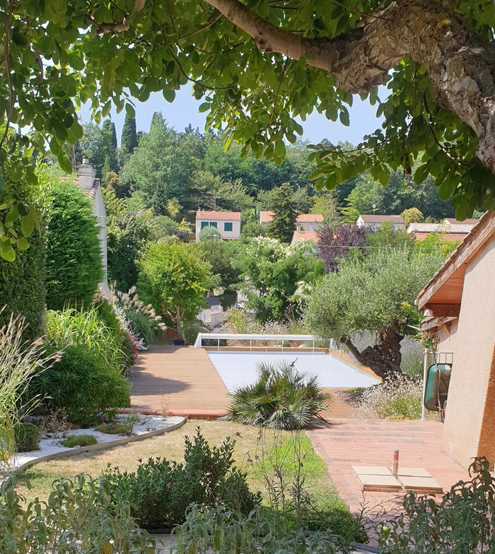 Vue d'un jardin luxuriant avec piscine couverte et terrasse en bois, entourée de verdure et de maisons en arrière-plan sous un ciel clair.
