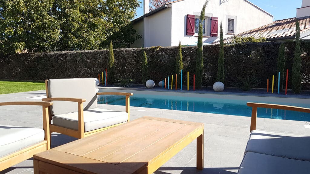 Mobilier de jardin en bois et coussins gris près d'une piscine bordée de galets, poteaux colorés et cyprès devant une haie.
