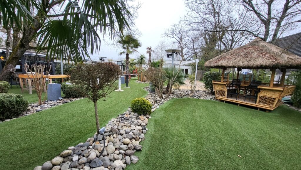 Jardin extérieur avec pelouse synthétique verte, chemin de galets, palmiers et un kiosque tiki en bambou pour se détendre.