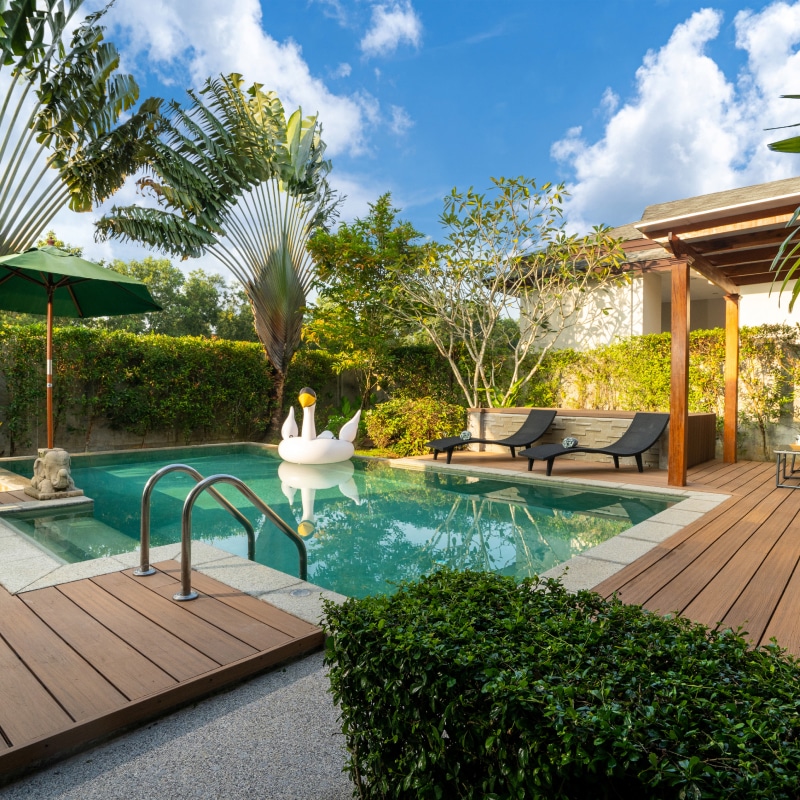 Piscine privée luxueuse dans jardin tropical, avec flamant gonflable blanc, chaises longues et parasol vert sous un ciel bleu.
