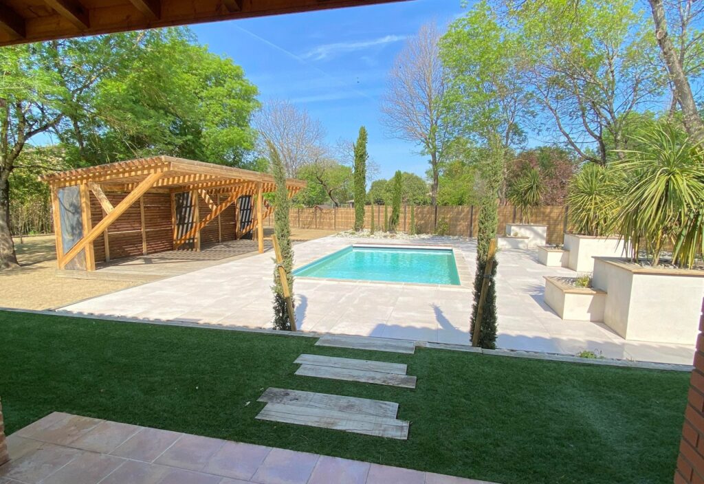 Piscine moderne et pergola en bois dans un jardin avec pelouse artificielle, dalles claires et plantes méditerranéennes sous un ciel bleu.