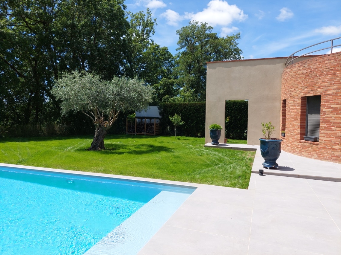 Piscine moderne aux margelles claires jouxtant une terrasse en dalles, jardin verdoyant avec un olivier centenaire et une architecture contemporaine en briques rouges et enduit beige.