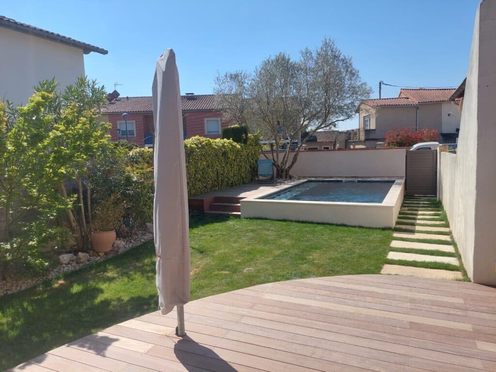 Jardin ensoleillé avec terrasse en bois, piscine rectangulaire enterrée, pelouse verte, parasol houssé et maison à l'arrière-plan.