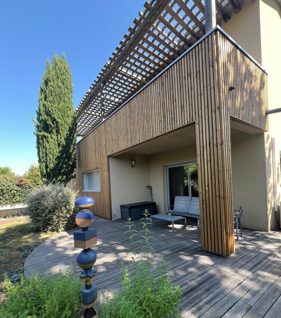 Terrasse en bois avec pergola, bardage vertical et totem artistique devant une maison moderne ensoleillée.