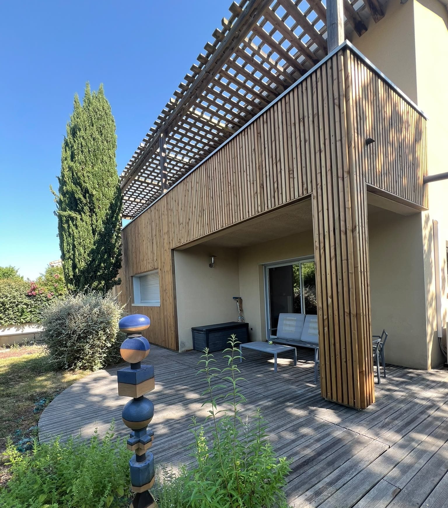 Maison moderne avec terrasse en bois, pergola et jardin Terrasse en bois avec pergola, bardage vertical et totem artistique devant une maison moderne ensoleillée.