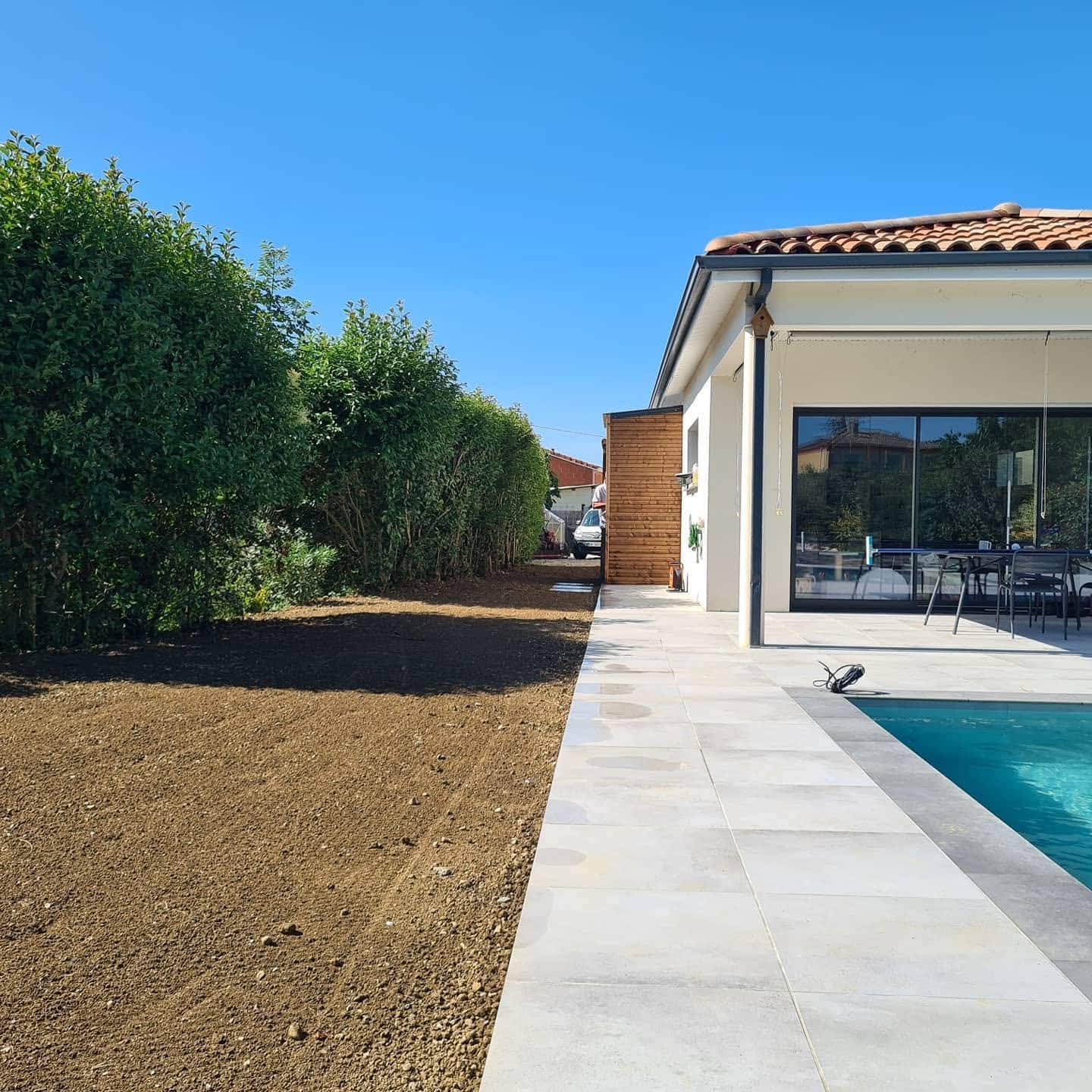Piscine, terrasse dallée et sol préparé pour gazon devant une haie sous un ciel bleu.