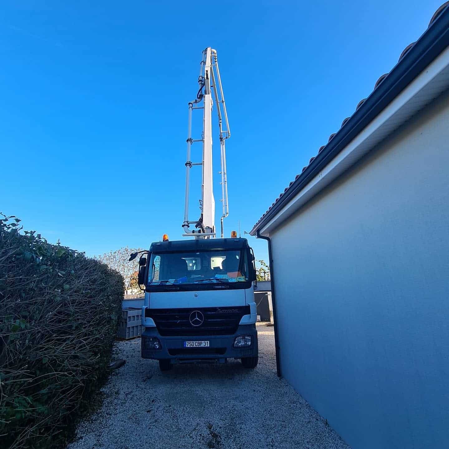 Pompe à béton Mercedes Actros 750 CBP 31, bras levé entre maison et haie sur gravier, ciel bleu.