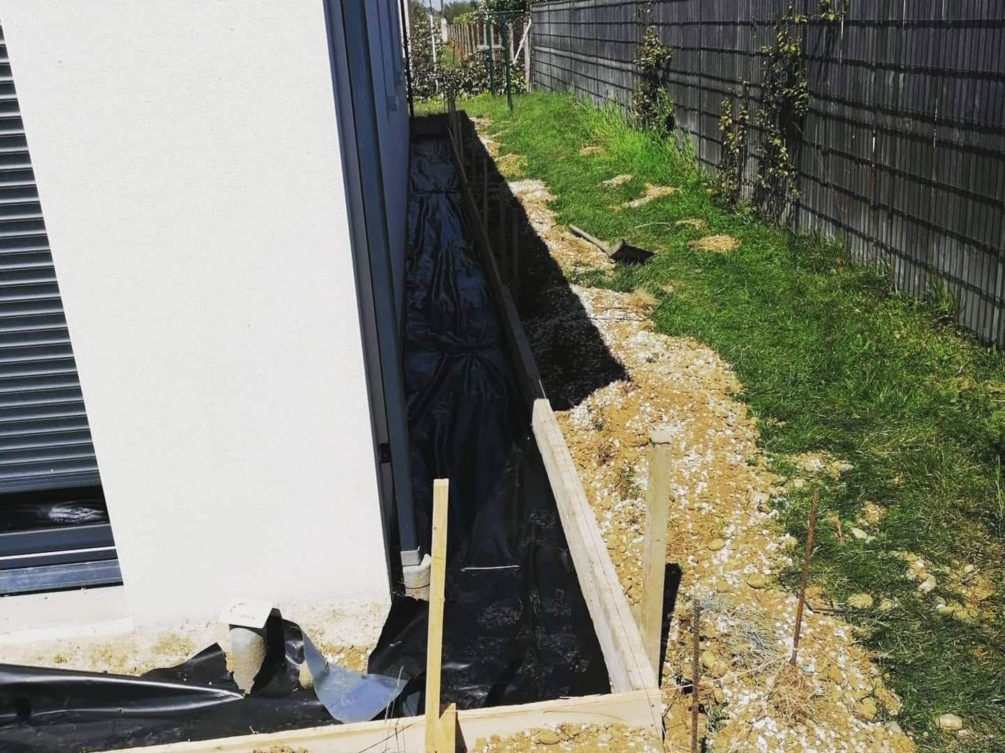 Pose de membrane d'étanchéité noire et coffrage bois pour drainage de fondation le long d'un mur blanc.
