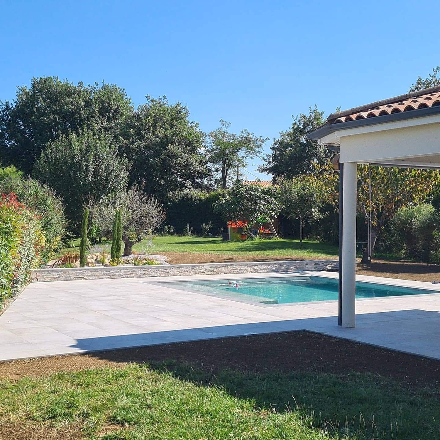 Piscine moderne avec terrasse en carrelage clair dans un jardin arboré sous un ciel bleu.