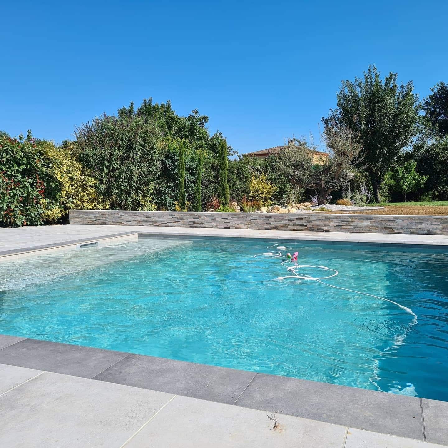 Piscine rectangulaire moderne avec eau bleue, terrasse grise, muret en pierre et jardin verdoyant sous un ciel pur.