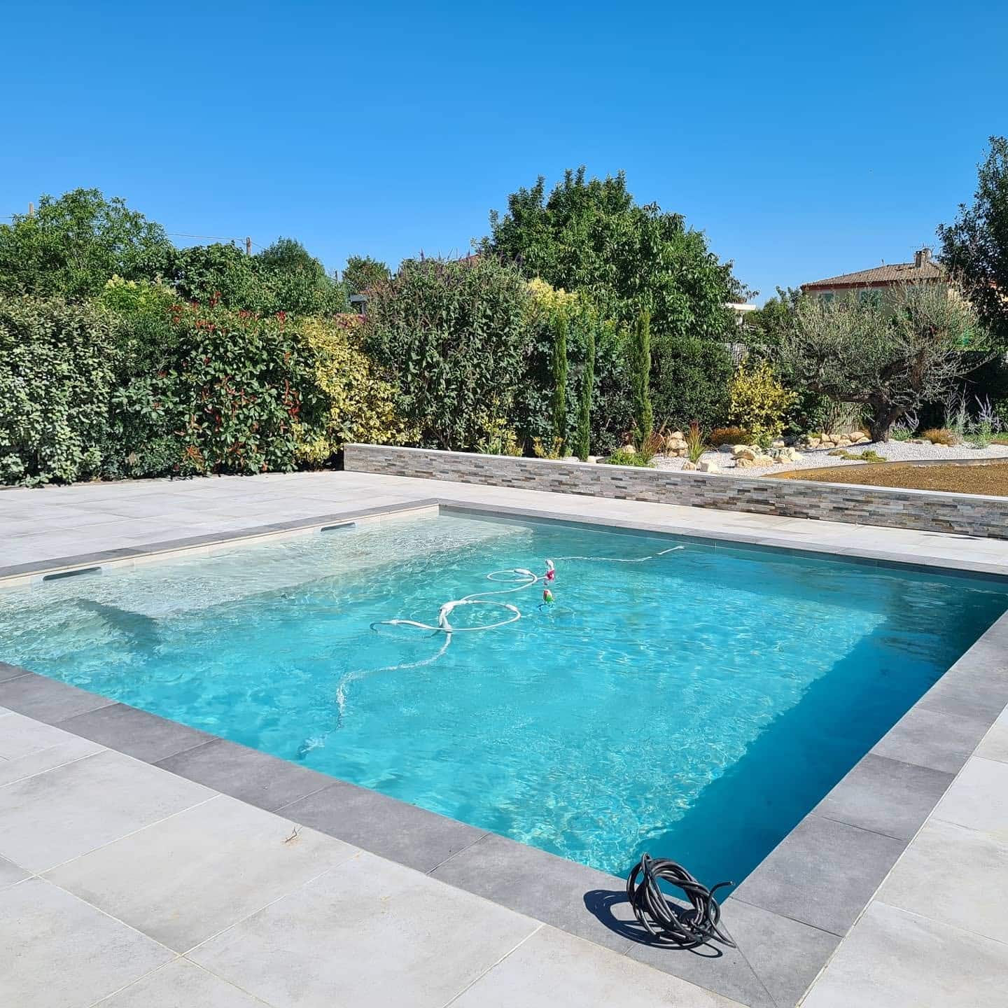 Piscine rectangulaire, eau turquoise, terrasse en dalles grises et jardin luxuriant sous ciel bleu.