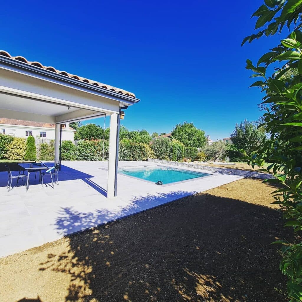 Terrasse moderne avec piscine rectangulaire, pergola blanche et jardin paysager sous un ciel bleu.