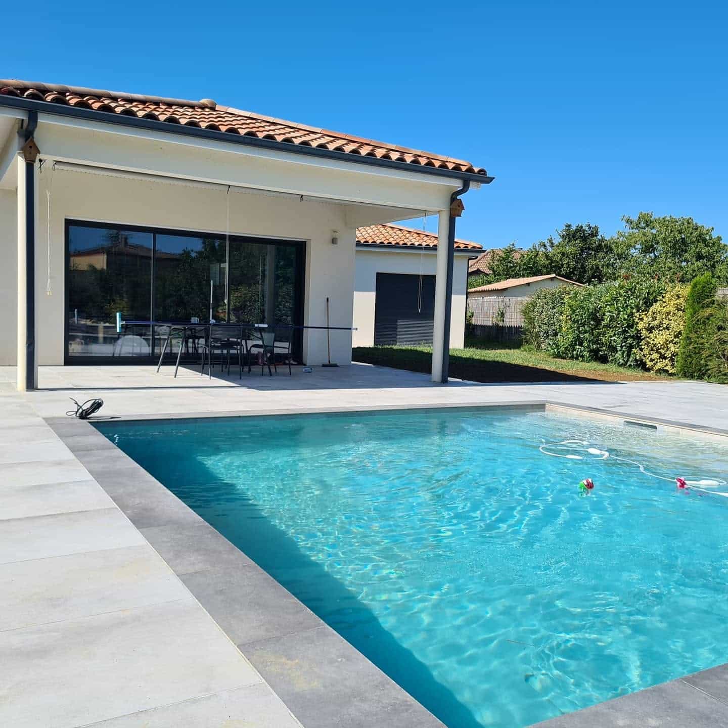 Villa moderne avec piscine aux eaux turquoise, terrasse en dalles claires et toit en tuiles sous un ciel bleu.