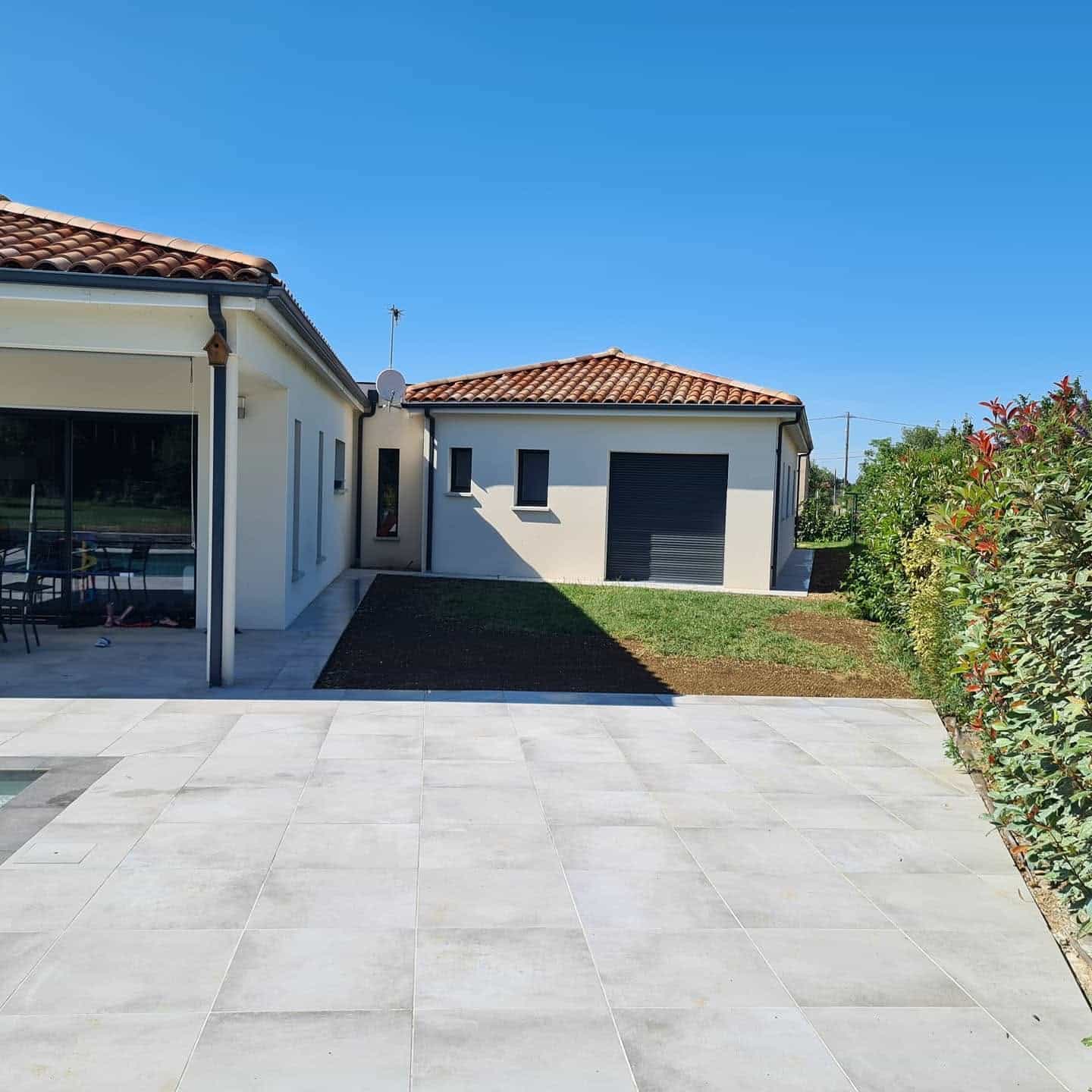 Terrasse carrelée grise devant une maison moderne à toit de tuiles sous un ciel bleu dégagé.