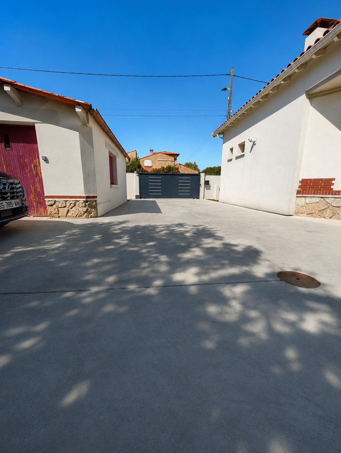 Aménagement Allée de Maison avec Portail Gris Moderne Allée en béton entre maisons blanches menant à un portail gris anthracite sous un ciel bleu ensoleillé.