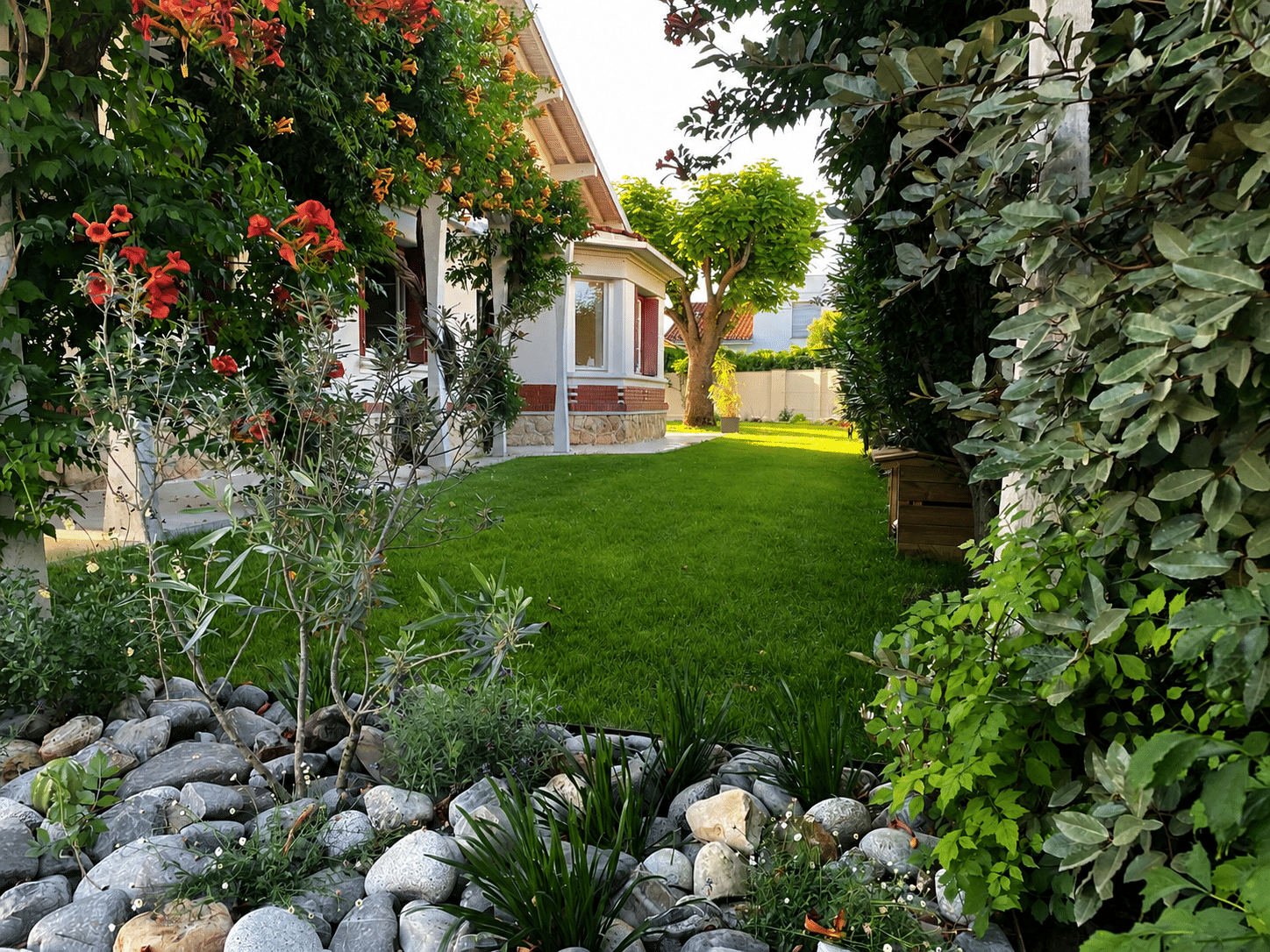 Jardin fleuri avec pelouse verte, fleurs de bignone, bordure de galets et maison blanche.