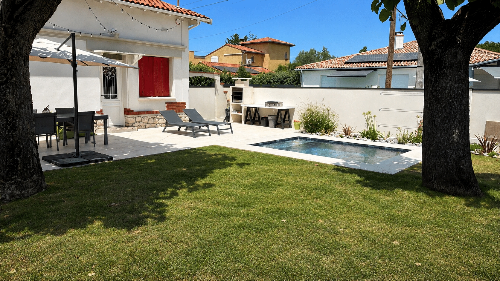 Jardin avec petite piscine, terrasse, transats et maison aux volets rouges sous le soleil.