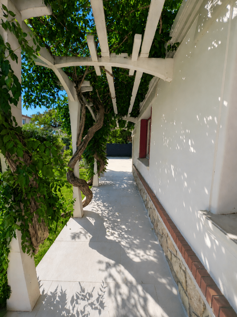 Pergola blanche avec vigne grimpante ombrageant une terrasse carrelée longeant un mur blanc au soleil.