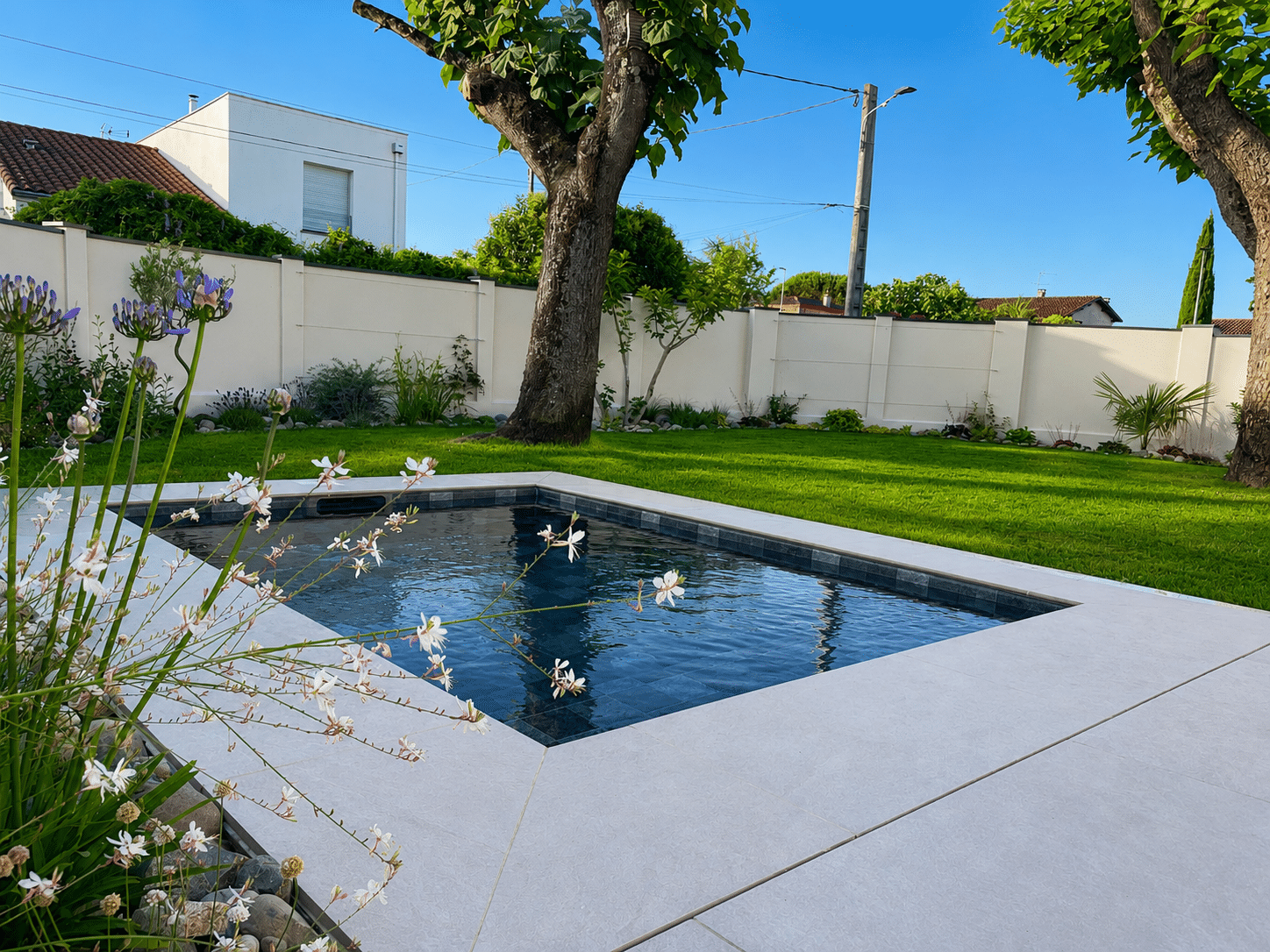 Petite piscine design avec margelles claires dans un jardin verdoyant et fleuri sous un ciel bleu dégagé.